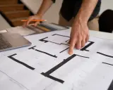 Dłoń wskazująca na plan mieszkania, obok laptop i próbki materiałów.