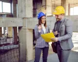 Architekci w kaskach rozmawiają na budowie, trzymając żółty folder.