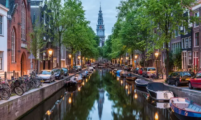 Kanał w Amsterdamie z łodziami, rowerami i wieżą kościelną w tle.